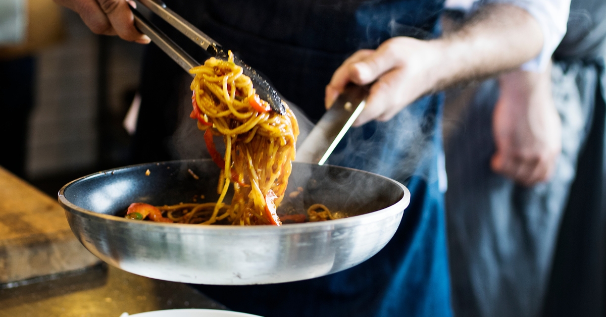 La imagen captura a un chef en acción, cocinando en una cocina profesional. Se le ve levantando una porción de espaguetis humeantes de un sartén metálico con unas pinzas, preparando la comida para ser servida. La imagen transmite la esencia de la cocina en vivo, mostrando la habilidad del chef y la calidad del plato en preparación.